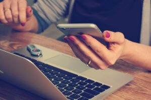 ノートパソコンの前でスマートフォンを操作する女性の手元 - デジタルデバイスに囲まれた仕事環境