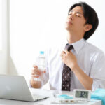 オフィスで水分補給をしながら暑さをしのぐビジネスマン。デスクには32℃を示す温度計と冷却タオルが置かれている