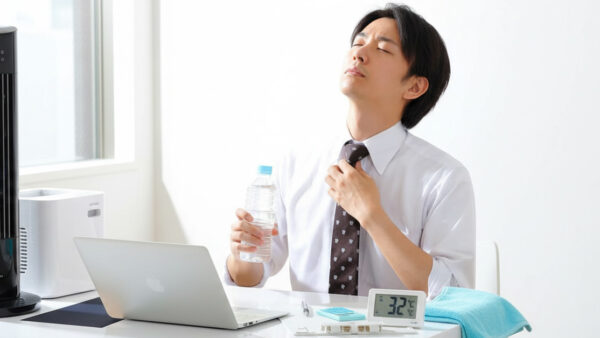 オフィスで水分補給をしながら暑さをしのぐビジネスマン。デスクには32℃を示す温度計と冷却タオルが置かれている