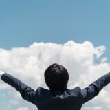 青空と雲を背景に両腕を広げるビジネスマン