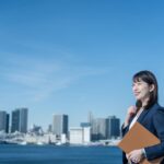 ビジネスウーマンが海辺で書類を持ち、都市風景を背景に立つ
