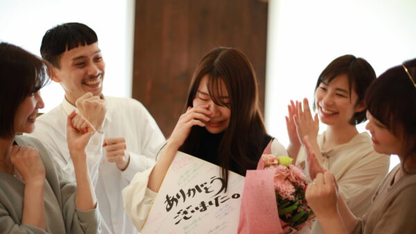 花束と寄せ書きを持つ女性社員と、拍手で送り出す同僚たち。オフボーディング（退職者の送り出し）のイメージ