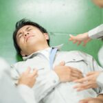 作業服を着た男性が倒れ、同僚が駆け寄る様子。労働災害発生時のイメージ